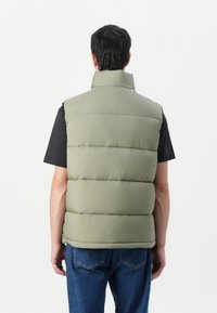 Puffer vest in muted olive green, featuring a high collar and horizontal stitching. Worn over a black t-shirt with visible denim jeans.
