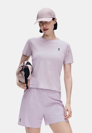 Frau trägt ein hellviolettes Sport-T-Shirt, Shorts und eine Kappe und hält eine passende Jacke vor einem weißen Hintergrund.