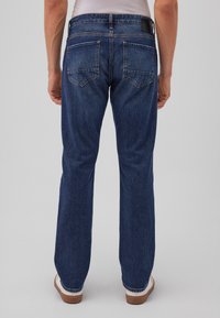 Hombre vestido con jeans de mezclilla azul rectos, calcetines blancos y zapatos marrones, de pie con los brazos relajados a los lados frente a un fondo gris liso.
