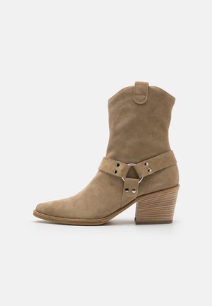 Beige suede ankle boot with a pointed toe, a stacked heel, and a decorative strap with circular hardware detail.