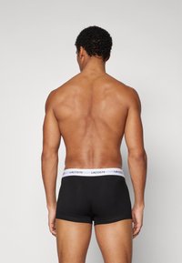 Schwarze Herrenboxershorts mit einem weißen Bund, der das Lacoste-Logo trägt, von hinten vor einem neutralen Hintergrund gezeigt.