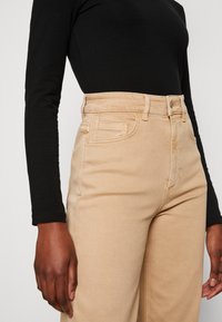 Jeans beige de cintura alta con bolsillos frontales y costuras en contraste, combinados con una blusa negra de manga larga. Textura de tela suave.