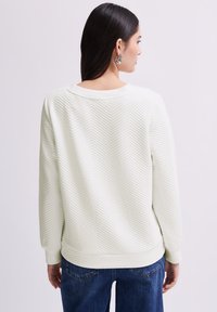 Maglione verde chiaro con texture caratterizzato da un motivo a spina di pesce, polsini e vita a coste, abbinato a jeans blu scuro.