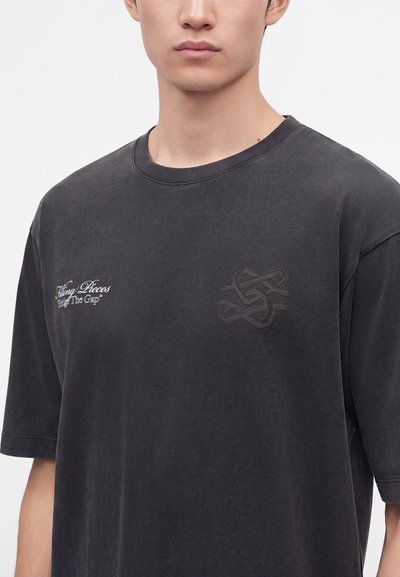 Νέο άτομο που φοράει ξεθωριασμένο μαύρο T-shirt με μικρό λευκό κείμενο στο αριστερό στήθος και διακριτικό σκούρο κεντητό λογότυπο στο δεξί στήθος.