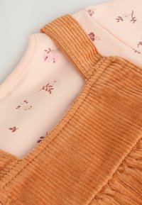 Salopette en velours côtelé orange brûlé avec des bretelles striées, associée à une chemise à manches longues à motif floral rose clair. Texture de tissu douce.