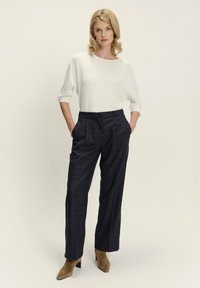 Weißes, geripptes Oberteil mit drei Viertelärmeln, kombiniert mit weiten Hosen im dunkelgrauen Karomuster und braunen Wildlederabsätzen. Schlichtes, modernes Design.