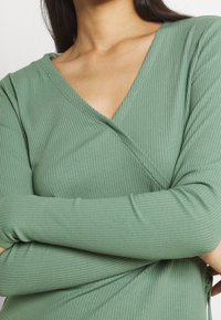 Zielony prążkowany top z długim rękawem, z dekoltem w kształcie litery V i skrzyżowanym detalem. Posiada miękką, teksturowaną tkaninę oraz dopasowany krój.