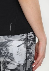 Svart atletisk topp med "FLASHDRY"-logotyp, kombinerad med grå mönstrade leggings med blommiga och randiga motiv, hand vilande på höften.