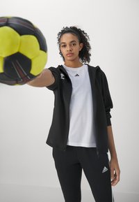 Ung kvinde i sportstøj holder en gul og sort håndbold fremadrettet, vendt mod kameraet med neutralt ansigtsudtryk mod en ensfarvet baggrund.