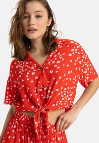Punapuku lyhythihainen crop-top, jossa on edessä solmu ja napitus. Pukussa on pieniä valkoisia kukkakuosia kankaassa, joka on teksturoitua.