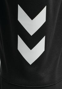 Czarne spodnie sportowe z teksturowanej tkaniny, z dużymi białymi wzorami w kształcie chevronu po bokach. Prosty design z dopasowaną sylwetką.