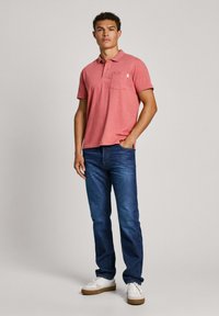 Camiseta polo roja con un bolsillo, combinada con jeans azules. El modelo lleva zapatillas blancas con acentos beige. Silueta simple y casual.