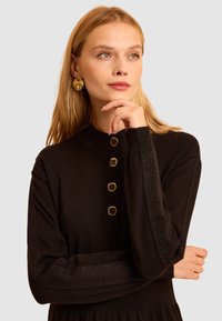 Czarna sukienka z długim rękawem, z wysokim dekoltem, złotymi guzikami oraz błyszczącymi, teksturowanymi akcentami na rękawach. Gładka tkanina z subtelnym połyskiem.