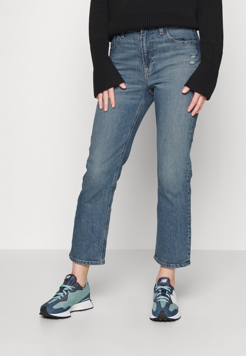 Personne portant un jean bleu taille haute, un pull noir avec des manches cloche et des baskets New Balance bleu-vert, debout les jambes écartées.