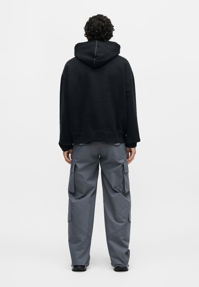 Άτομο με σγουρά μαλλιά που φοράει μαύρο φούτερ με κουκούλα και γκρι παντελόνι cargo, στέκεται και φαίνεται από πίσω, με ένα απλό φόντο.