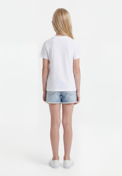 Παιδί με μακριά ξανθά μαλλιά που φοράει απλό λευκό μπλουζάκι, ανοιχτόχρωμο τζιν σορτς και άσπρα παπούτσια, στέκεται γυρισμένο με την πλάτη σε λευκό φόντο.