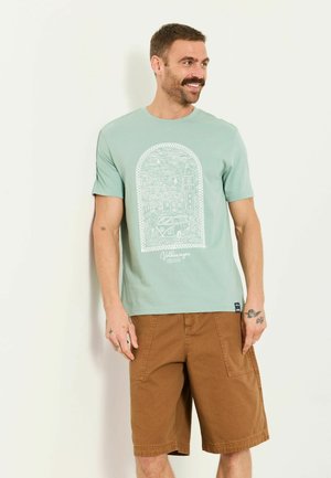 Lächelnder Mann trägt ein hellgrünes Volkswagen-Grafik-T-Shirt und braune knielange Shorts, steht vor einer einfarbigen hellen Wand.