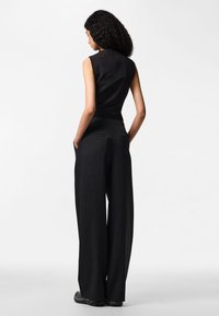 Sieviete ar cirtainiem matiem stāv ar muguru, valkājot melnu bezrokas topu un platas melnas bikses ar smalkām svītrām, rokās ieliktas kabatās.