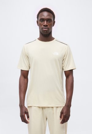 Młody czarnoskóry mężczyzna stojący przodem, ubrany w beżową koszulkę North Face z krótkim rękawem i pasujące spodnie na jednolitym białym tle.