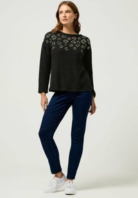 Schwarzes Langarmshirt mit einem Blumenmuster aus Perlen auf der oberen Vorderseite, kombiniert mit dunkelblauen, eng anliegenden Hosen und weißen Turnschuhen.