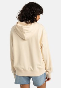 Felpa con cappuccio color crema, dal taglio morbido e rilassato, con spalle scese e polsini a coste, abbinata a pantaloni corti di jeans.