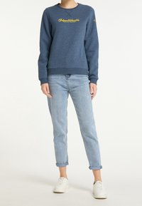 Sudadera de cuello redondo azul hecha de una mezcla de algodón, con texto amarillo bordado. Combinada con jeans de puños enrollados de color azul claro y zapatillas blancas.