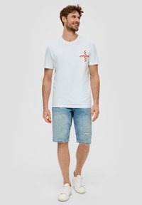 Camisa de algodón blanca con un gráfico pixelado rojo y blanco en el lado izquierdo del pecho, combinada con pantalones cortos de mezclilla azul claro y zapatillas blancas.