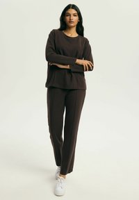 Brązowa bluzka z długim rękawem, okrągłym dekoltem i bocznymi rozcięciami, w zestawie z dopasowanymi spodniami o prostych nogawkach. Modelka ma na sobie białe trampki.