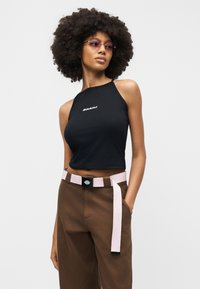 Γυναίκα με μεγάλο, σγουρό μαλλί που φοράει μοβ γυαλιά ηλίου, μαύρο crop top χωρίς μανίκια με λευκό λογότυπο, καφέ παντελόνι και ανοιχτό ροζ ζώνη.