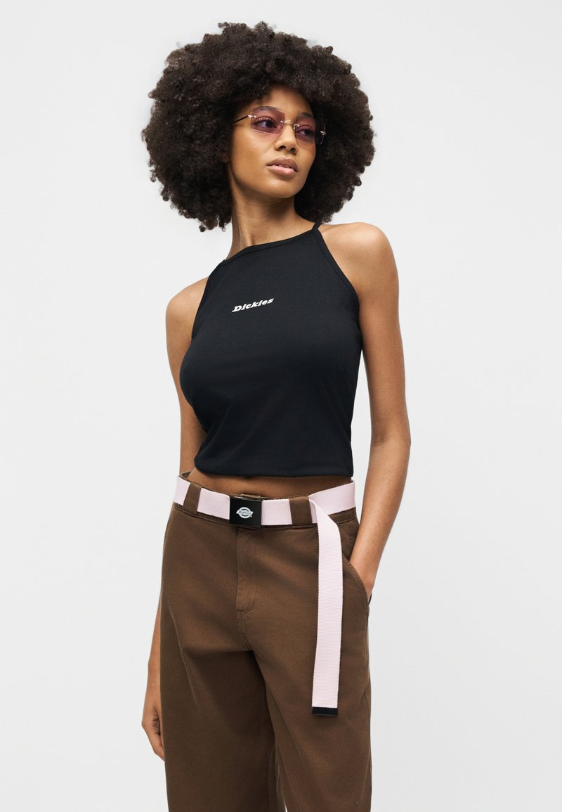 Woman with large curly hair wearing purple sunglasses, sleeveless black crop top with white logo, brown pants, and light pink belt.