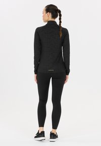 Vrouw met gevlochten haar, gekleed in een zwart sporttop met lange mouwen en leggings, staande met de rug naar de camera op een witte achtergrond.