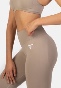 Béžové legíny s vysokým pásom vyrobené z hladkej, elastickej tkaniny s diskrétnym logom na stehne a textúrovaným detailom pásu.