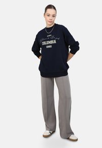 Felpa oversize blu navy con testo e logo decorativi bianchi, abbinata a pantaloni larghi grigi chiari con dettagli di cuciture verticali.