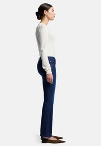 Una modella è in profilo mentre indossa un maglione aderente color crema e jeans a zampa d'elefante blu scuro, completati da eleganti scarpe leopardate a punta.