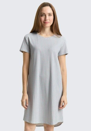 Woman with straight brown hair wearing a short-sleeve, knee-length, light-colored dress with a small geometric pattern against a plain background.