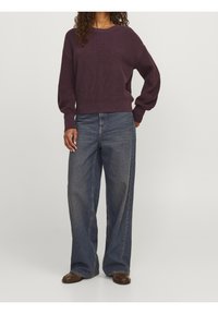 Maglione lavorato a maglia viola con ampio scollo a girocollo, maniche lunghe e orlo a coste, abbinato a jeans in denim blu dalla vestibilità ampia e stivaletti marroni.