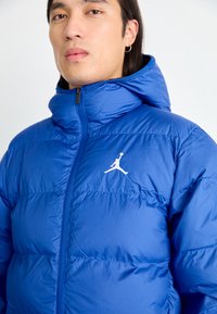 Blå pufferjakke med hette, laget av skinnende materiale. Har en frontglidelås og en hvit Jordan-logo på brystet. Teksturert, quiltet design.