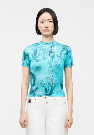 Femme aux cheveux noirs et lisses portant un haut turquoise ajusté à manches courtes avec des motifs bleus tourbillonnants, et un pantalon blanc taille haute à boutons dorés.