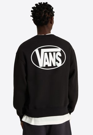Personne aux cheveux foncés coiffés en petites torsades porte un sweat-shirt noir avec un grand logo Vans blanc dans le dos, sur une chemise blanche.