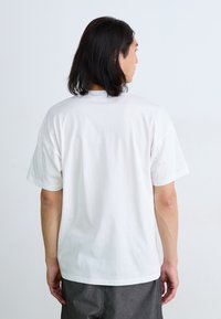 T-shirt en coton blanc avec un col rond et des manches courtes, présentant un dos uni et une coupe décontractée, porté avec un pantalon gris. Pas de motifs ni de graphiques.