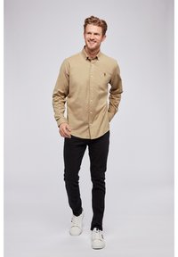 Beige gombos ing gallérral, hosszú ujjakkal és logós részlettel, fekete farmerrel és fehér sportcipővel párosítva. Sima textúra.