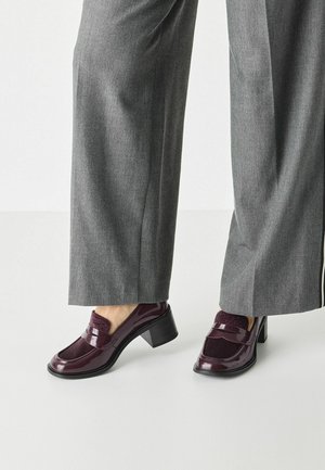 Burgunderrøde laklæder loafers med hæl og teksturerede detaljer, kombineret med grå bukser med vide ben, der fremhæver et slankt design.