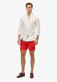 Chemise en lin blanche, shorts de bain rouges avec des accents noirs et du texte, et des chaussures de bateau marron. Les shorts sont dotés d'une taille élastique avec cordon de serrage.