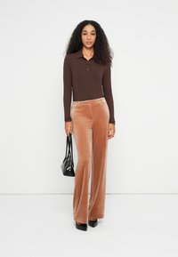 Mujer con cabello rizado que lleva una camiseta de manga larga de canalé marrón, pantalones anchos de terciopelo beige, tacones negros y sostiene un bolso negro.