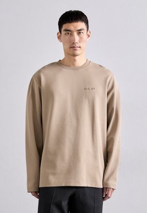 Beige long-sleeve t-shirt made from soft cotton. Features a crew neck and subtle embroidered detail on the chest. Loose fit, minimal design.