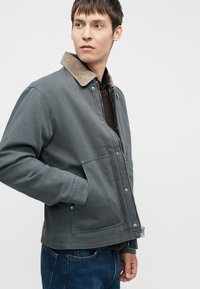 Grijze canvas jas met een bruine corduroy kraag, een rits aan de voorkant en drukknoopsluitingen. Bevat twee zijzakken en heeft een ontspannen pasvorm.