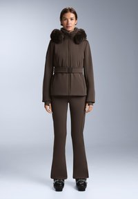 Kobieta ubrana w brązową zimową kurtkę z paskiem i futrzanym kołnierzem, dopasowane spodnie w tym samym kolorze oraz czarne śniegowce, stojąca na jednolitym tle.