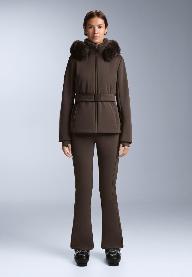 Ženska v rjavi zimskij jakni z rjavim pasom in krznenim ovratnikom, s hlačami, ki se prilegajo, in črnimi snežnimi škornji, stoji pred enobarvnim ozadjem.