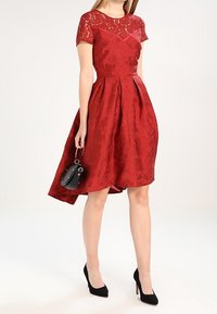 Robe en dentelle rouge avec des manches courtes, corsage ajusté, jupe plissée et ourlet asymétrique. Assortie à des escarpins noirs et un petit sac à main.