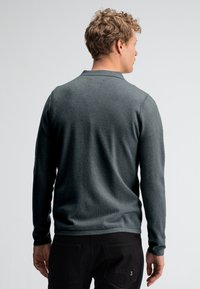 Dunkelgrüner Langarm-Polo-Pullover. Weicher Stoff, gerippter Kragen, taillierte Passform. Getragen mit schwarzen Hosen, minimaler Markenaufschrift auf dem Stoff.